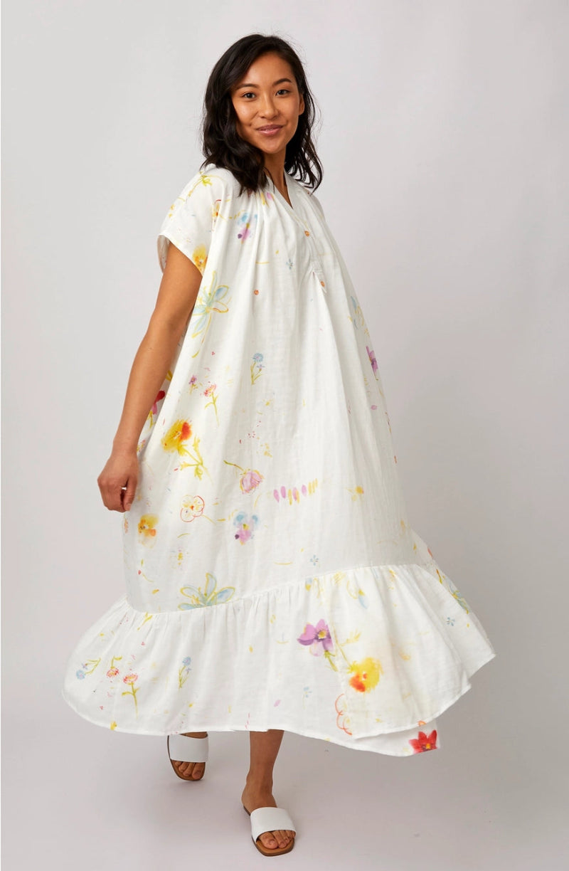 A woman modeling a white short-sleeve maxi dress with a colorful watercolor floral print, featuring a tiered ruffle hem, set against a plain light gray studio background.