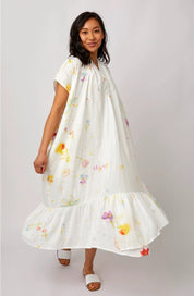 A woman modeling a white short-sleeve maxi dress with a colorful watercolor floral print, featuring a tiered ruffle hem, set against a plain light gray studio background.