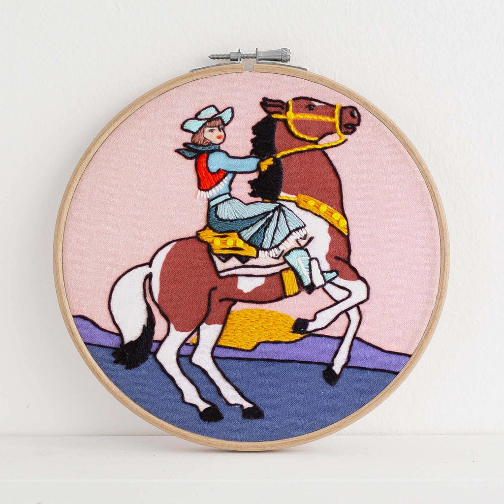 Embroidery hoop with a colorful depiction of a person riding a horse on a pink background