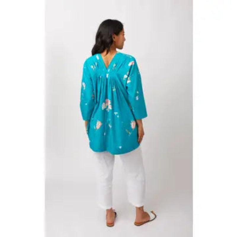 A woman shown from the back wearing a turquoise blue floral tunic top with white pants and sandals, standing against a light gray studio background.