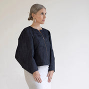 person wearing a dark denim jacket and white pants against a plain background