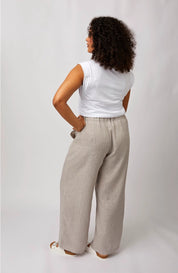 A woman with curly dark hair is shown from the back wearing wide-leg light beige trousers, a white sleeveless top, and white slide sandals against a light gray studio background.