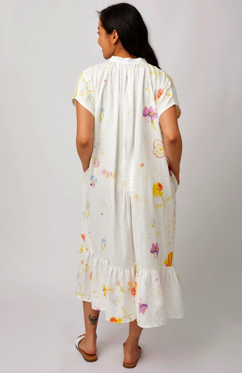 A woman viewed from behind wearing a white short-sleeve midi dress with a colorful floral print, paired with brown flat sandals, against a light gray studio background.