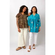 Two women modeling casual tunic tops and white pants against a plain light gray studio background. The woman on the left wears a brown patterned short-sleeve tunic with cream pants, and the woman on the right wears a turquoise floral long-sleeve tunic with white pants.
