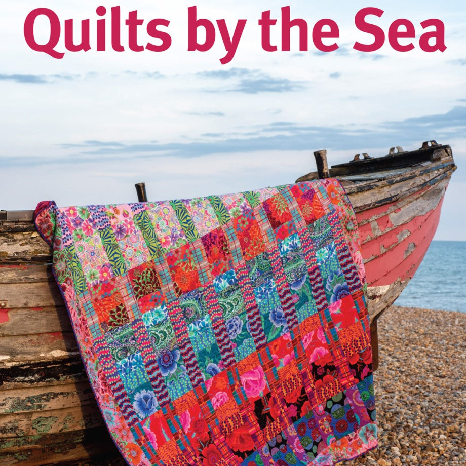 Colorful quilt on a wooden boat by the sea with 'Kaffe Fassett's Quilts by the Sea' text.