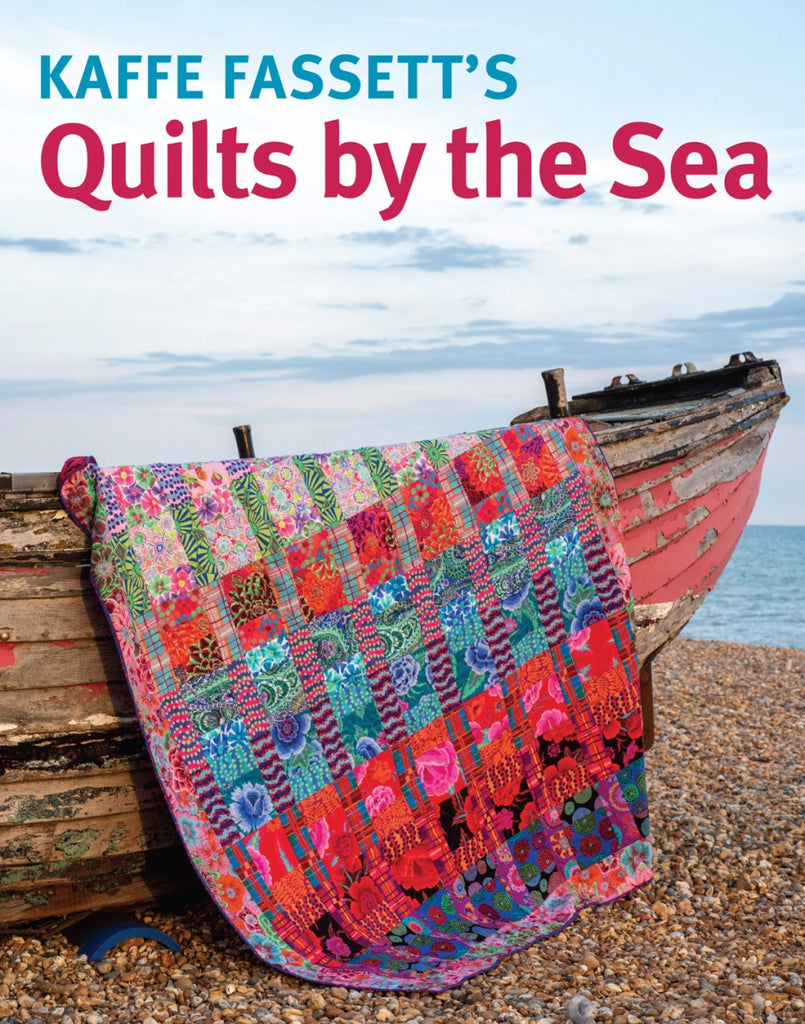 Colorful quilt on a wooden boat by the sea with 'Kaffe Fassett's Quilts by the Sea' text.