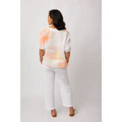 A woman shown from the back wearing a white short-sleeve top with a soft peach and yellow tie-dye pattern, paired with white wide-leg pants and brown sandals, against a light gray studio background.