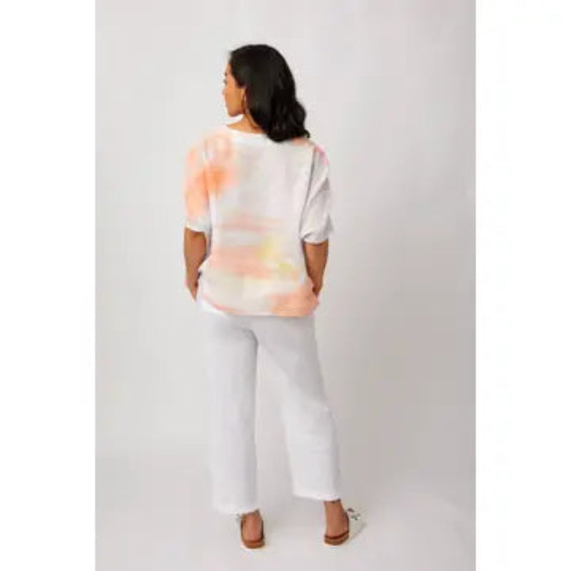 A woman shown from the back wearing a white short-sleeve top with a soft peach and yellow tie-dye pattern, paired with white wide-leg pants and brown sandals, against a light gray studio background.