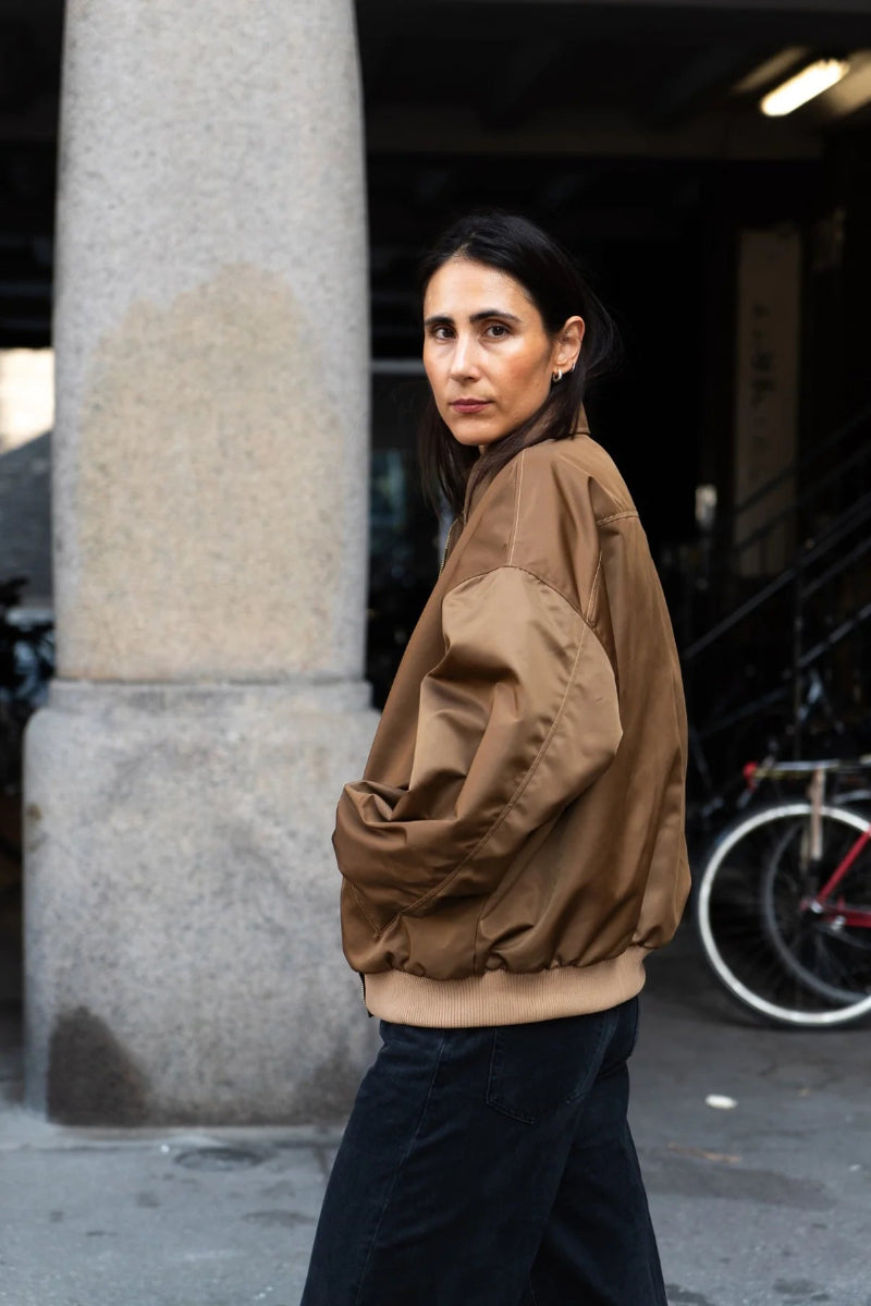 Person wearing a brown jacket standing next to a column on a street.