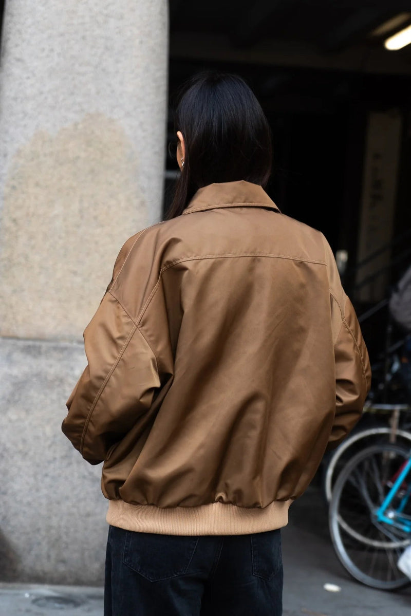 Person wearing a brown jacket standing next to a stone column with a bicycle in the background.