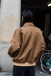 Person wearing a brown jacket standing next to a stone column with a bicycle in the background.