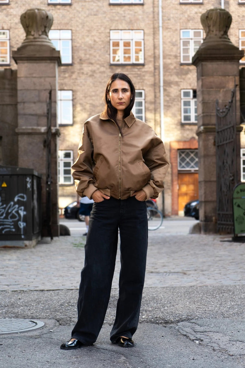 Person wearing a brown jacket and black pants standing in an urban setting with buildings and parked cars in the background.