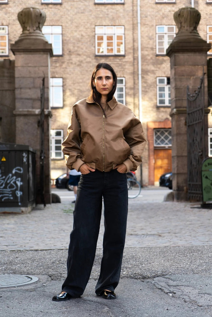 Person wearing a brown jacket and black pants standing in an urban setting with buildings and parked cars in the background.