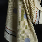 A close-up of a cream-colored fabric garment, likely a sari or scarf, featuring a fine gold dot pattern, large light blue-gray circular motifs, and a decorative border with red, black, and white stripes.