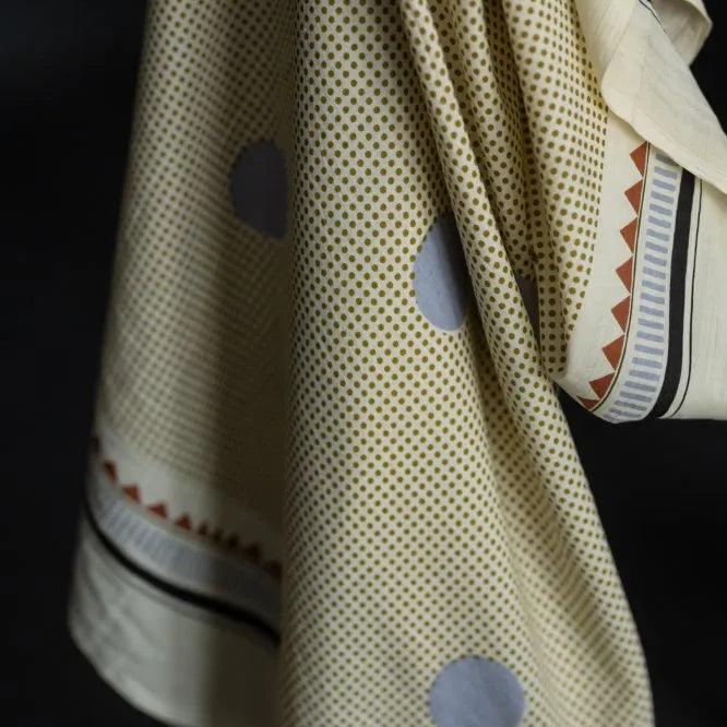A close-up of a cream-colored fabric garment, likely a sari or scarf, featuring a fine gold dot pattern, large light blue-gray circular motifs, and a decorative border with red, black, and white stripes.