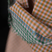 A close-up of folded fabric with a small gingham check pattern in green, orange, and white, layered over a beige textured fabric against a dark background.