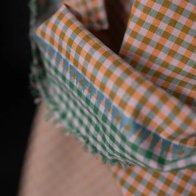 A close-up of folded fabric with a small gingham check pattern in green, orange, and white, layered over a beige textured fabric against a dark background.