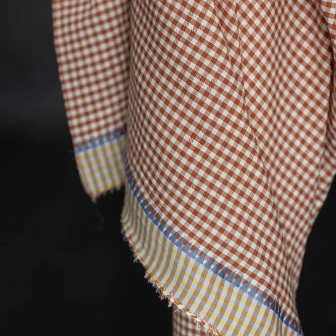 A close-up of a gingham fabric with a brown and white checkered pattern and a contrasting striped border in yellow, blue, and white, draped against a dark background.
