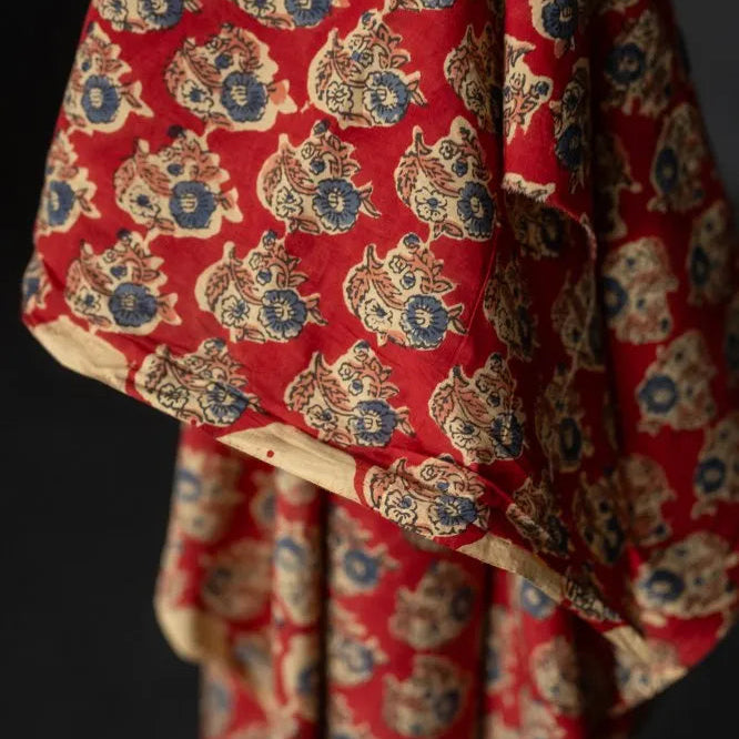 A close-up of a draped red fabric with an all-over block-printed pattern of stylized faces in beige and blue, set against a dark background.
