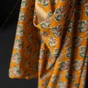 A close-up of a draped mustard yellow fabric with an all-over floral block print in muted green, pink, and cream, set against a dark background.