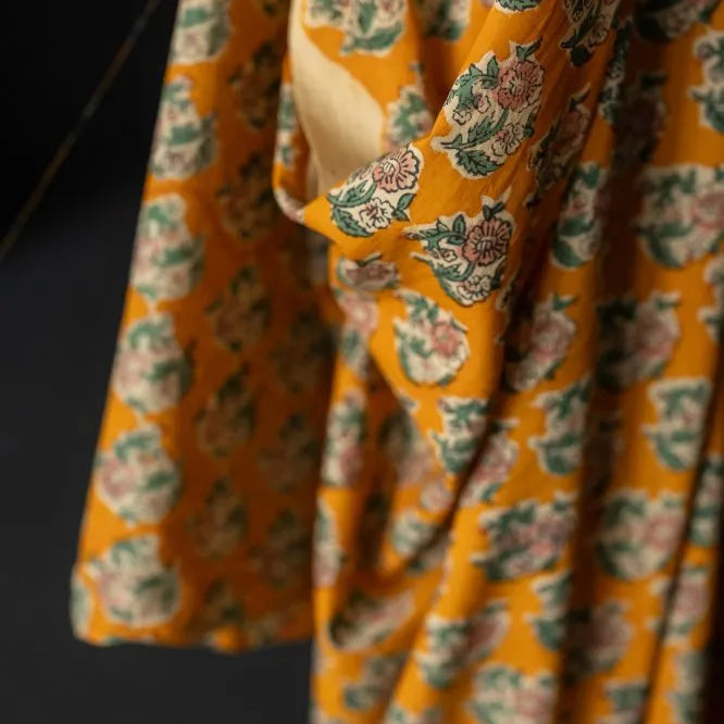 A close-up of a draped mustard yellow fabric with an all-over floral block print in muted green, pink, and cream, set against a dark background.
