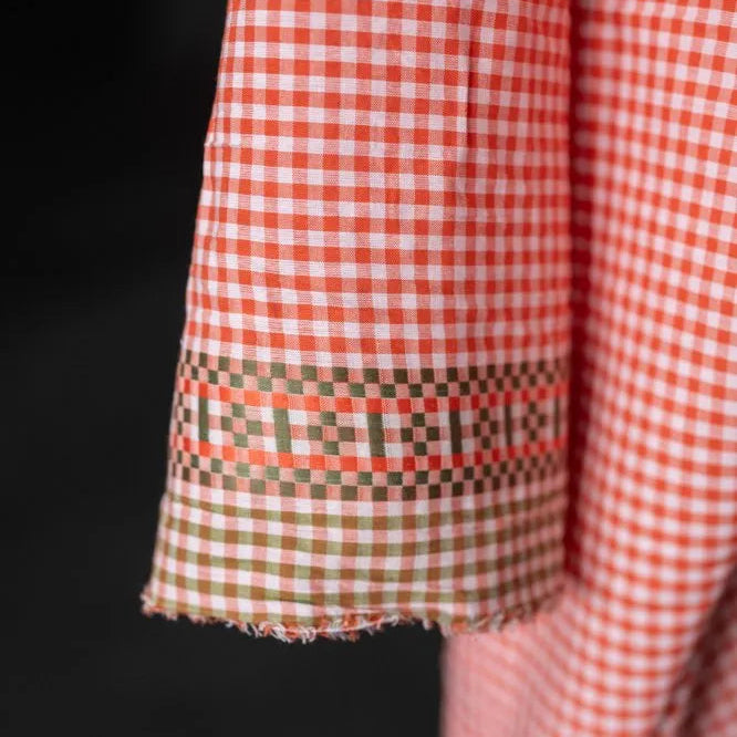 Close-up of a red and white gingham fabric sleeve with a decorative band of green, orange, and red checkered pattern near the cuff, set against a dark background.