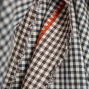 A close-up of a fringed checkered fabric with a brown and white gingham pattern and a blue and white gingham pattern, showing a small orange accent stripe.