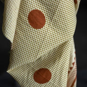 A close-up of a cream-colored fabric with a fine brown polka dot pattern and two large rust-brown circular patches, set against a dark background.