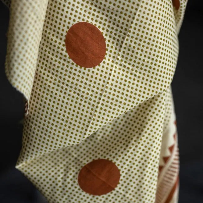 A close-up of a cream-colored fabric with a fine brown polka dot pattern and two large rust-brown circular patches, set against a dark background.