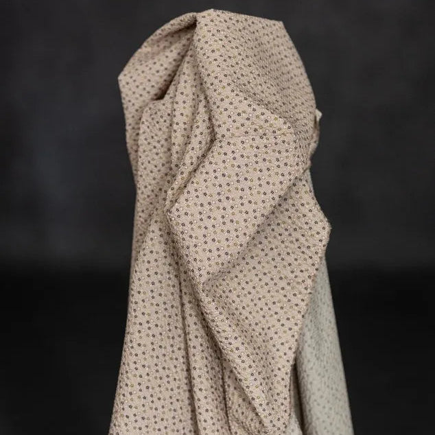 A draped piece of light beige fabric with a small, evenly spaced brown geometric pattern, shown against a dark, textured background.