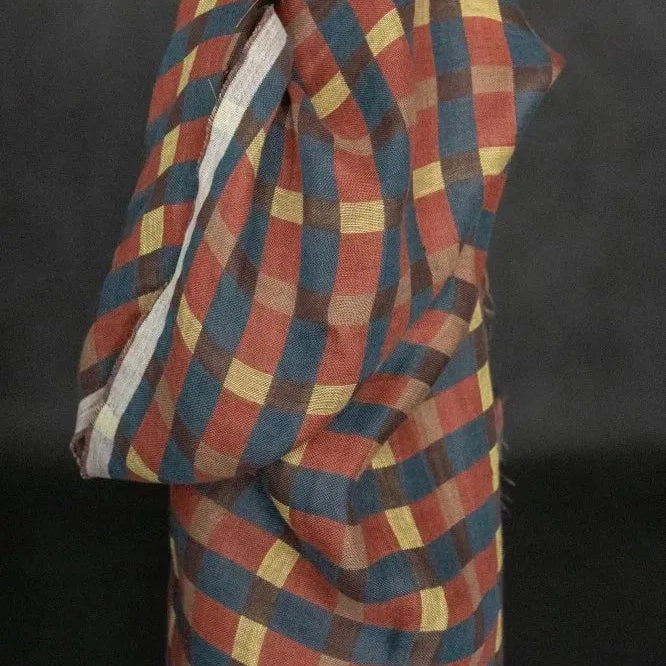 A close-up of a draped fabric or textile with a diagonal plaid/check pattern in muted red, blue, yellow, and brown tones against a dark background.