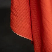 A close-up of a textured red-orange fabric with a fine stripe, hanging against a dark background.