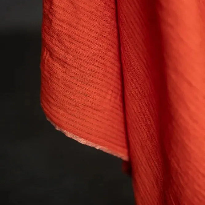 A close-up of a textured red-orange fabric with a fine stripe, hanging against a dark background.