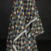 A draped fabric item, likely a shawl or scarf, featuring a geometric diamond pattern in olive green, brown, navy blue, and cream, with a contrasting light gray border, set against a dark background.