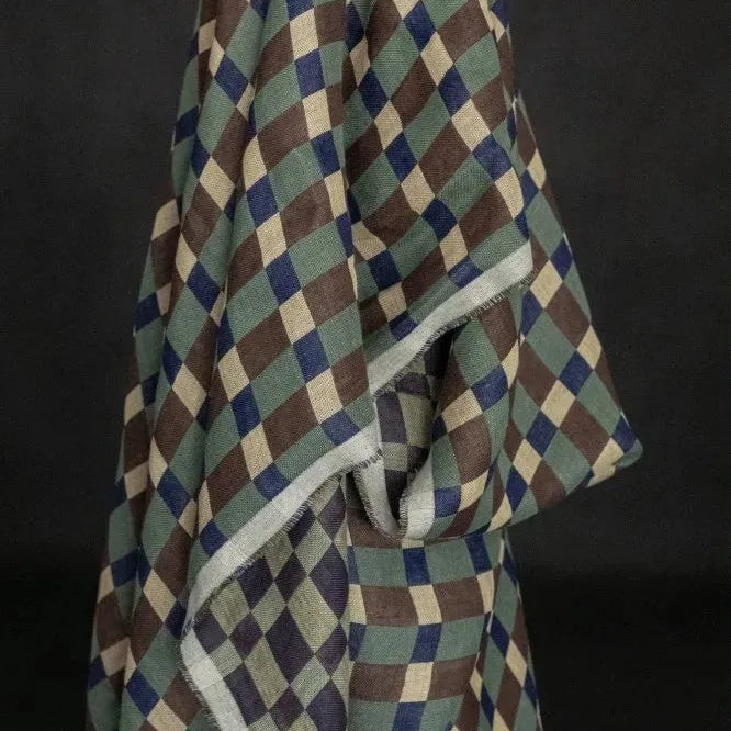 A draped fabric item, likely a shawl or scarf, featuring a geometric diamond pattern in olive green, brown, navy blue, and cream, with a contrasting light gray border, set against a dark background.