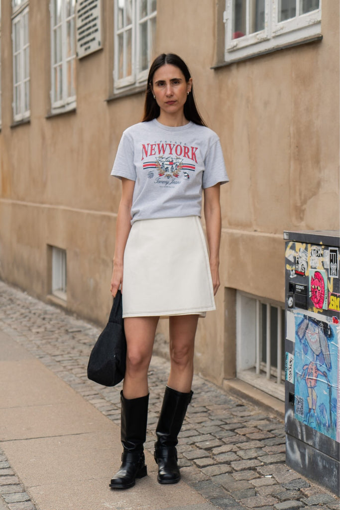 Person wearing a 'New York' t-shirt and white skirt standing on a street.