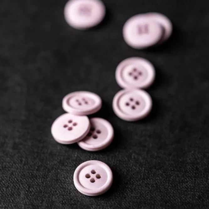 Several small round four-hole buttons in a pale pink or dusty mauve color scattered on a dark textured fabric background.