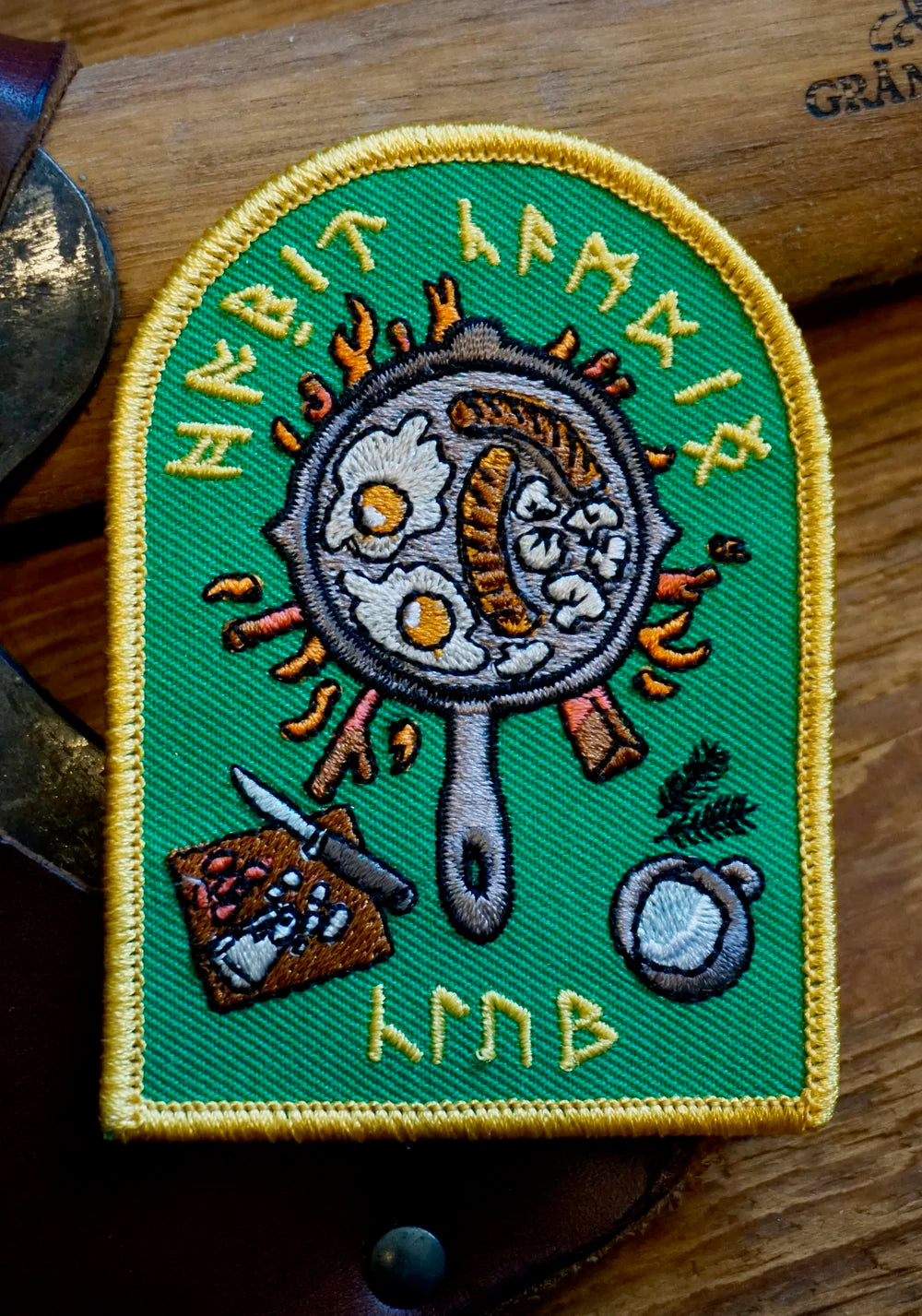 Embroidered patch with a frying pan, eggs, and runes on a green background.