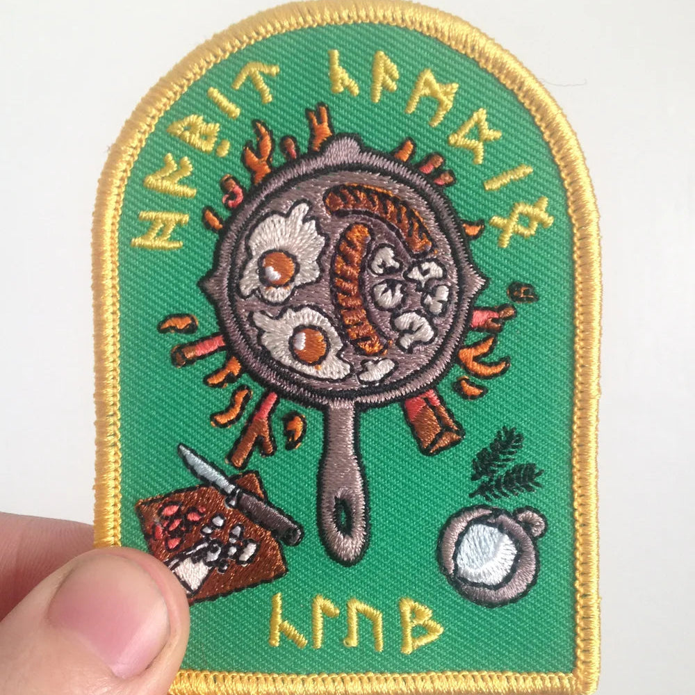 Embroidered patch with a frying pan design and runes on a green background, held by a hand.