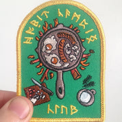 Embroidered patch with a frying pan design and runes on a green background, held by a hand.