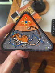 Triangular embroidered patch with a mouse design on a wooden surface