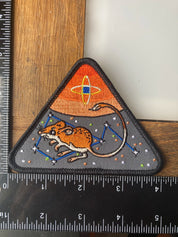 Triangular patch with a mouse design and space theme, next to a ruler for scale.