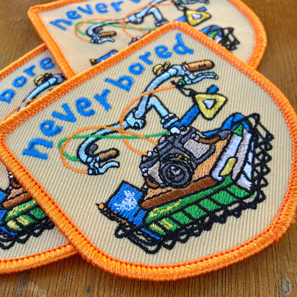 Two embroidered patches with a bike design and 'never bored' text on a wooden surface.