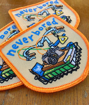 Two embroidered patches with a bike design and 'never bored' text on a wooden surface.