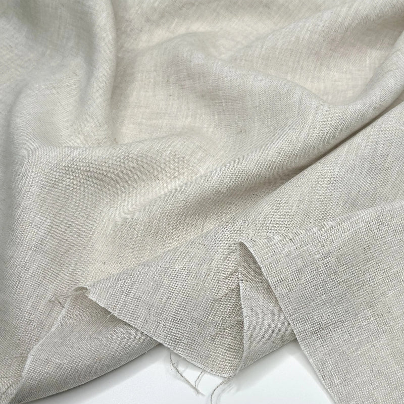A close-up of draped light beige linen fabric with a visible woven texture and frayed edges, laid over a white surface.