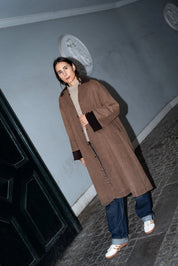 Person in a brown coat standing on a street with a light blue wall and dark door in the background.