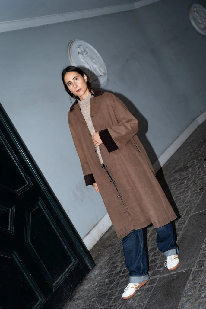 Person in a brown coat standing on a street with a light blue wall and dark door in the background.