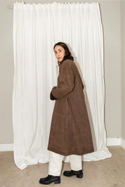 Person wearing a brown coat standing in front of white curtains.