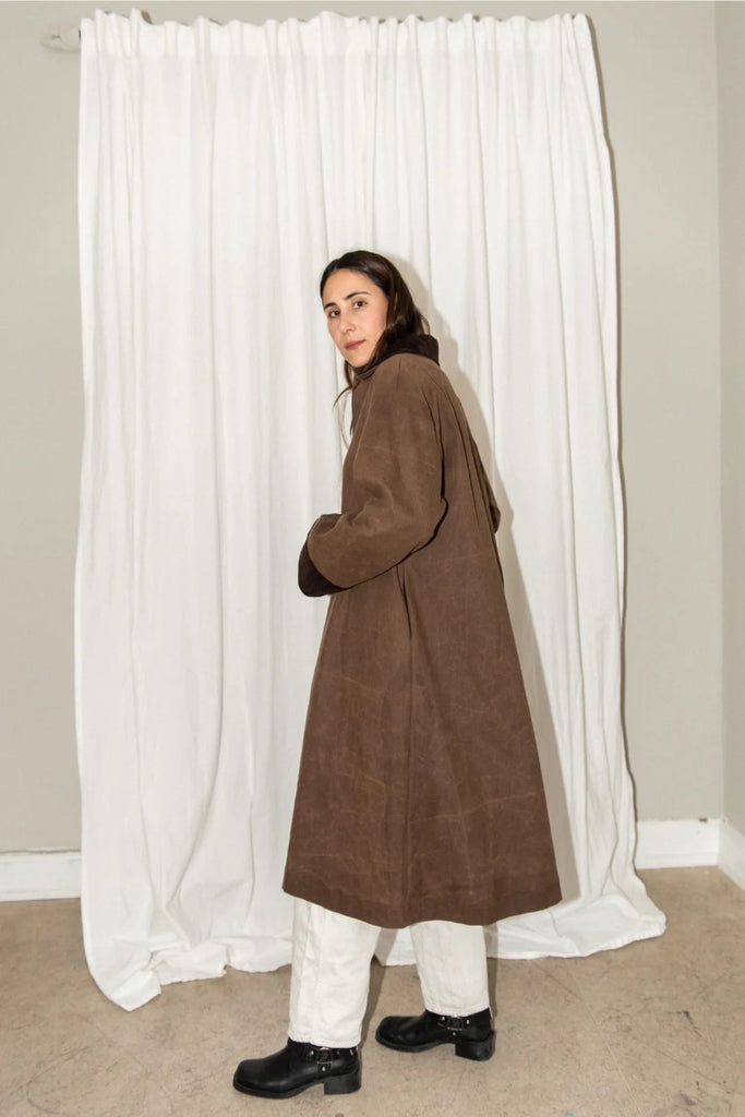 Person wearing a brown coat standing in front of white curtains.