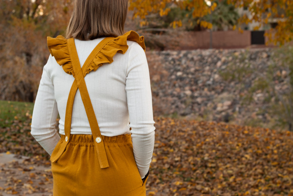 Person wearing mustard yellow overalls with ruffled straps in an outdoor setting with autumn leaves.